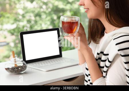 Junge Frau, die heissen Tee genießt, während sie ferngesteuert auf einem Laptop mit leerem Bildschirm arbeitet, gemütliches Heimbüro Stockfoto