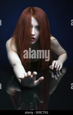Schöne erschreckende junge Frau mit Ingwerhaar über Spiegelspiegel auf blauem Hintergrund Stockfoto