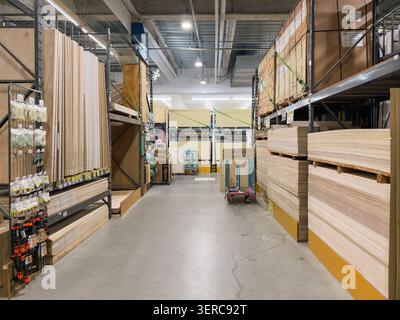 Ein geräumiger Gang in einem japanischen Baumarkt zeigt Holzpaneele, die auf beiden Seiten von hohen Regalen gestapelt sind, mit einem Einkaufswagen und weiteren Produkten, die sich in den Hintergrund erstrecken. Stockfoto