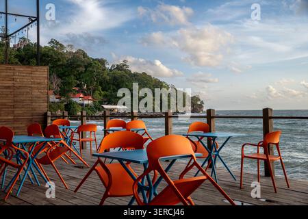 Café-Terrasse am Strand mit leuchtenden orangefarbenen Stühlen und blauen Tischen mit Blick auf das tropische Meer und die Hügel. Stockfoto