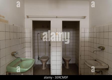 Innenraum eines verlassenen institutionellen Badezimmers mit zwei Toiletten, alten Podest-Toiletten mit hohen Spülkästen und einem grünen Keramikwaschbecken auf der Th Stockfoto