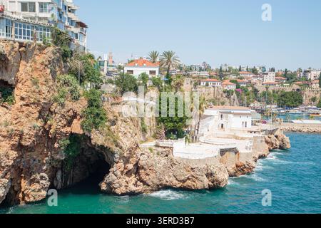 Hohe Kalksteinklippen mit natürlichen Grotten und alte osmanische Häuser mit Ziegeldächern über dem klaren Wasser des alten Kaleici Hafens. lan an der Küste im Sommer Stockfoto