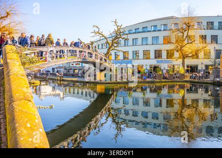 LONDON, 2. MÄRZ 2025: Menschenmassen versammeln sich auf der eisernen Fußbrücke über den Regents Canal in Camden, London. Die Szene umfasst Reflexionen im Wasser und Gebäude im Hintergrund an einem sonnigen Tag. Stockfoto