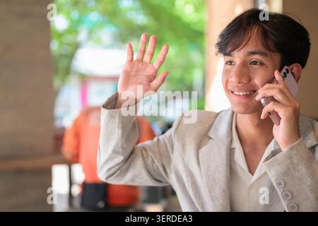 Authentisches horizontales Lifestyle-Porträt eines jungen, nicht-binären Thai-Isan-Profis oder -Studenten, der lächelt und hallo winkt, während er am Telefon spricht. Setzen Sie Sich Stockfoto
