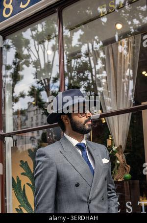 Ein stilvoller, bärtiger Mann mit Anzug, klassischem blauem Fedorahut und Sonnenbrille steht vor einem eleganten Ladenfenster in London. Stockfoto