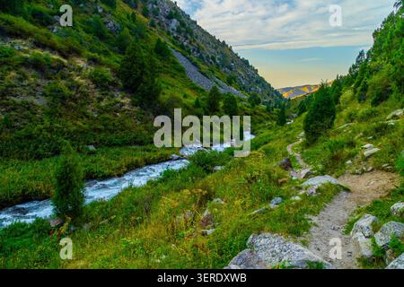 Die malerische Bergtallandschaft bietet einen sich windenden Fluss, der durch üppiges Grün in Kirgisistan fließt. Ein unbefestigter Wanderweg führt den felsigen Hang hinauf, Stockfoto