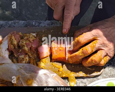 Nahaufnahme der Hände, die mariniertes Fleisch für das Grillen im Freien vorbereiten. Rohe Fleischstücke, die mit Gewürz und Marinade beschichtet sind, werden geschnitten und eingefädelt Stockfoto