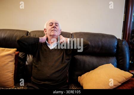 Ein älterer Mann sitzt auf einem Sofa und massiert seinen Hals wegen Schmerzen und Steifheit Stockfoto