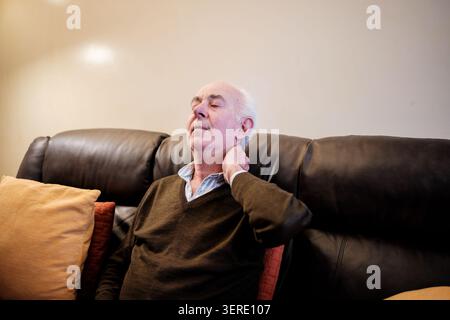 Ein älterer Mann leidet unter Nackenschmerzen, während er auf einem braunen Ledersofa sitzt Stockfoto