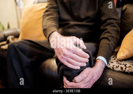 Ein älterer Mann, der an Knieschmerzen leidet, sitzt auf einem Sofa in seinem Wohnzimmer Stockfoto
