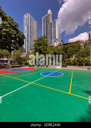 Moderne Wolkenkratzer ragen hinter einem lebendigen, frisch gestrichenen Basketballplatz in einem geschäftigen Stadtpark empor und symbolisieren eine dynamische Mischung aus Natur. Stockfoto