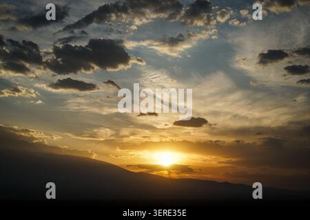 Eine leuchtende goldene Sonne taucht unter eine dunkle, silbrige Bergkette und beleuchtet den Horizont in warmen Tönen. Oben füllt sich ein dynamischer Himmel mit dunklem, weißem Himmel Stockfoto