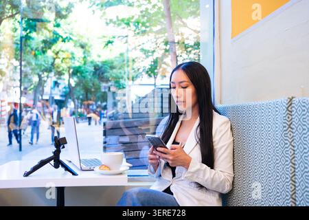 Frau, die sich auf das Smartphone konzentriert, von einem Café aus mit Laptop und Kaffee aus arbeitet Stockfoto