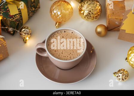 Das aufschäumende Kaffeegetränk in einer pastellfarbenen Tasse fängt das gemütliche Gefühl der Weihnachtszeit ein, umgeben von festlichen Goldverzierungen und Geschenkverpackungen Stockfoto