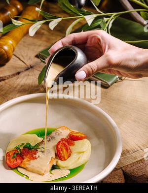 Eine Hand gießt eine cremige Soße über eine wunderschön plattierte Lachsschale, garniert mit Kirschtomaten und Kräutern. Die Szene spielt auf einem rustikalen Holztisch mit Grün im Hintergrund. Stockfoto