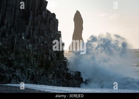 Der Strand von Reynisfjara zeigt mächtige Meereswellen, die auf dunkle Basaltsäulen mit einem fernen Meeresstapel unter hellem Himmel stürzen. Vik Island Stockfoto