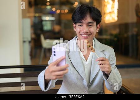 Authentisches horizontales Lifestyle-Porträt eines jungen nicht-binären Thai Isan Food Influencers oder Vloggers, der nachts ein Selfie macht. Einen pinkfarbenen Smartph hochhalten Stockfoto