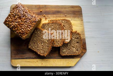 Ansicht von oben nach unten auf geschnittenem Vollkornbrot mit Sonnenblumenkernen, die auf rustikalem Holzschneidebrett auf einer hellen Tischplatte angeordnet sind, sichtbarer Krümelstab Stockfoto