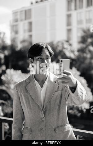 Vertikale mittlere Aufnahme eines jungen, nicht binären Geschlechts fließenden Thai Isan Geschäftsmannes, der einen eleganten lässigen grauen Blazer und ein weißes Hemd mit Kragen trägt, lächelnd Stockfoto
