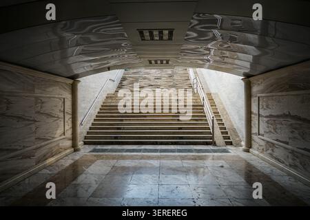 Eine breite, leere Marmortreppe erhebt sich in einem großen architektonischen Innenraum nach oben, mit kunstvollen Steinmauern und einem einzigartig reflektierenden, wellenförmigen cei Stockfoto