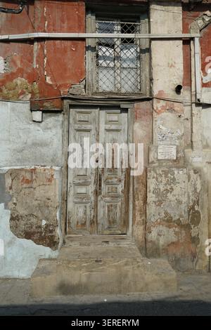 Eine alte, verwitterte Doppeltür aus Holz mit verblasster Farbe und ein Sicherheitsfenster darüber sind an einer verzweifelten Gebäudefassade zu sehen. Der Krumblin Stockfoto