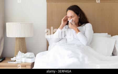 Kranke Frau niest in Gewebe, während sie im Bett sitzt und Erkältungssymptome zeigt Stockfoto