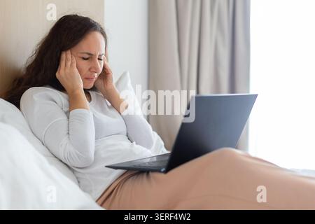 Frau, die im Bett mit Laptop ruht und an Kopfschmerzen leidet Stockfoto