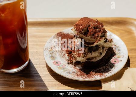 Köstliches Tiramisu-Dessert mit Kakaopulver und eiskaltem Americano-Kaffee auf einem Holztablett unter hellem Sonnenlicht Stockfoto