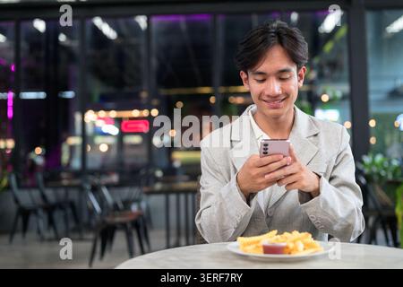 Horizontaler mittlerer Schuss eines jungen nicht binären Geschlechts fließenden Thai Isan Geschäftsmann, der einen eleganten lässigen grauen Blazer und ein weißes Hemd trägt. Auf einer Terrasse sitzen Stockfoto