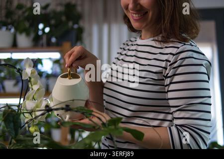 Lächelnde Frau mit einer stilvollen Gießkanne, die ihre schöne weiße phalaenopsis-Orchidee und verschiedene Zimmerpflanzen pflegt, die friedliche und erfüllende Atmosphäre genießen Stockfoto