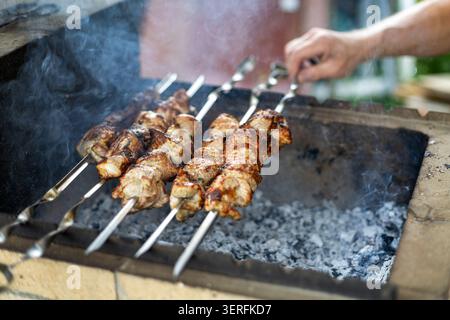 Gegrillte Fleischspieße, die über heißen Kohlen kochen. Gegrillte Fleischspieße kochen über rauchig heißen Kohlen, während eine Hand sie auf einem Grill im Freien dreht. Stockfoto