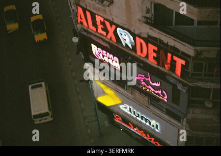 Ein Hochwinkel bei Nacht mit Blick von einem Fenster im oberen Stockwerk auf eine Straßenecke in Alexandria, Ägypten. Ein auffälliges rot beleuchtetes Schild für „ALEX D Stockfoto