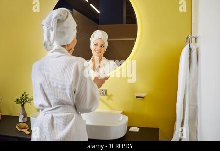 Frau mit Bademantel, Badetuch mit Turban und Blick auf den Spiegel im hellen, modernen Badezimmer Stockfoto