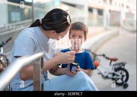 Eine Frau im Alter von 30 Jahren teilt einen freudigen Moment mit ihrem vierjährigen Sohn, während sie draußen Eis genießen. Die Szene fängt ihre Interaktion mit dem Fahrrad ein Stockfoto