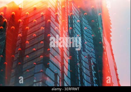 Künstlerische Doppelbelichtung eines modernen Glashochhauses in kräftigen Rot- und Blautönen mit analogem Filmlicht-Leck-Overlay, wodurch eine surreale Stadt entsteht Stockfoto