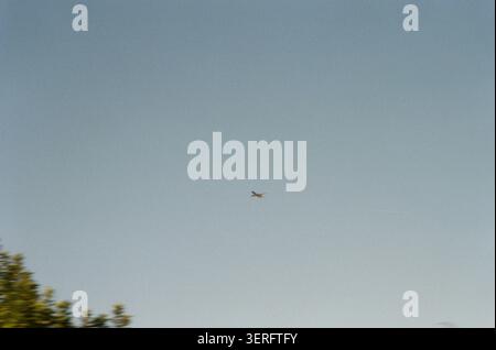 Ein weit entferntes kleines Flugzeug schwebt durch einen klaren, trüben blauen Himmel mit verschwommenen Baumkronen in der unteren Ecke. Aufgenommen auf Film mit einem nostalgischen, se Stockfoto