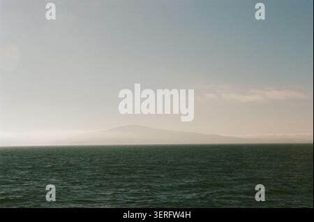 Das riesige, dunkelgrüne Meer erstreckt sich bis zu einem trüben Horizont, an dem ferne Berge durch den Nebel der Küste unter einem blassblauen Himmel auftauchen. Moody, atmosphärische Meereslandschaft Stockfoto