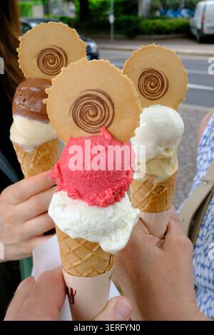 Drei Hände, die Gelato-Waffeln mit bunten Kugeln und spiralförmigen Waffelkeksen auf einer Straße der Stadt halten, zeigen ein lustiges Sommervergnügen, Freundschaft und Lässigkeit Stockfoto
