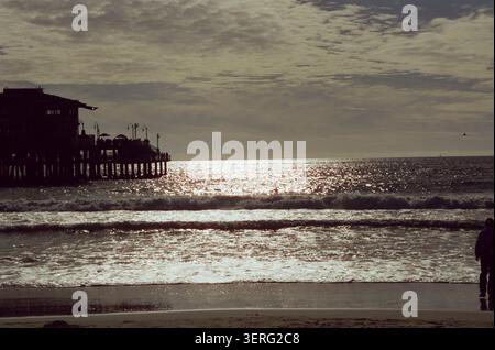 Dramatische Silhouette eines hölzernen Piers und Gebäudes vor einem glitzernden Meer bei Sonnenuntergang, mit rollenden Wellen und einem stimmungsvoll bewölkten Himmel. Stockfoto