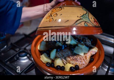Die Hand der Frau hebt den Deckel aus einem traditionellen marokkanischen Tagine-Topf und enthüllt Hühnchen, gehackte Kartoffeln und schwarze Oliven, die auf einem Gasherd kochen Stockfoto