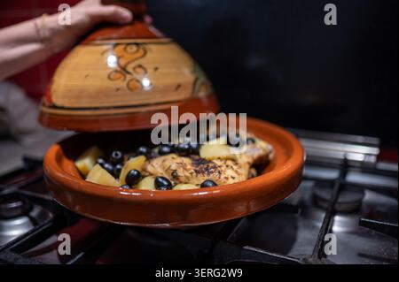 Person kocht traditionelle marokkanische Hühnertagine, kocht mit Kartoffeln und Oliven in einem Tontopf auf einem Gasherd und bereitet eine aromatische und herzhafte Zubereitung zu Stockfoto