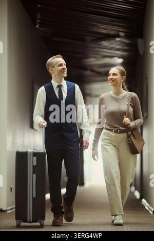 Vertikale Aufnahme einer kaukasischen Frau und eines männlichen Porträts, die mit Koffern durch den Hotelflur laufen, um dort Urlaub oder Geschäftsreise zu machen. Ideal für Reisebuchungen, Hospitality Marketing Stockfoto