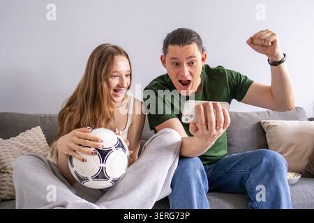 Aufgeregt Vater und Teenager-Tochter feiern das Fußballergebnis, während sie auf den Smartphone-Bildschirm schauen. Familie teilt emotionalen Sport-Moment, unterstützt Stockfoto
