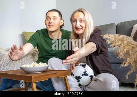 Paare reagieren emotional, während sie Fußballspiele mit Foodball und Popcorn zu Hause anschauen. Mann und Frau, die Sportsendungen verfolgen, unterstützen Stockfoto