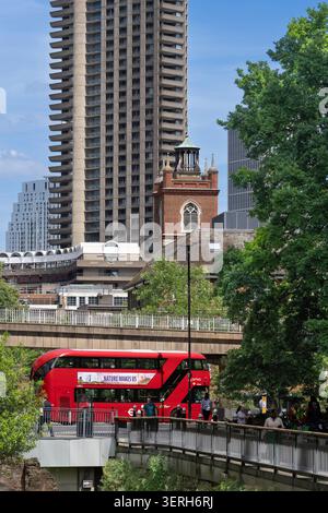 Die Leute pendeln an einem roten Bus vorbei am Lauderdale Tower und St Giles' Cripplegate im Barbican Estate in London für ihre täglichen Aktivitäten. Stockfoto