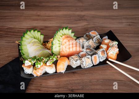 Sushi-Sortiment mit Brötchen, Sashimi und Nigiri auf schwarzem Teller gegen dunklem Holz. Fokus auf japanische kulinarische Kunst, gesunde Zubereitung von Speisen und Stockfoto