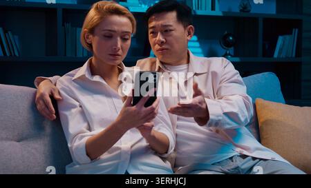 Multiethnische Paare sprechen mit dem Mobiltelefon die Bestellung hat abends zu Hause unterschiedliche Meinungen. Der asiatische Mann stimmt nicht mit dem Kaukasier überein Stockfoto