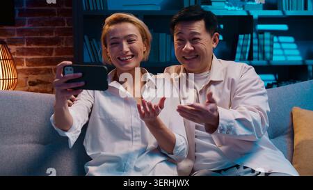 Glückliches fröhliches multiethnisches Paar lächelnd Videoanruf mit Handy-Chat im abendlichen Wohnzimmer. Optimistischer asiatischer Mann mit Familie Stockfoto
