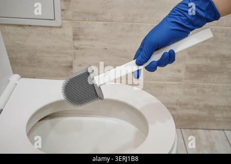 Person mit blauem Schutzhandschuh, der Silikon-Toilettenbürste hält, die moderne Toilette reinigt Stockfoto
