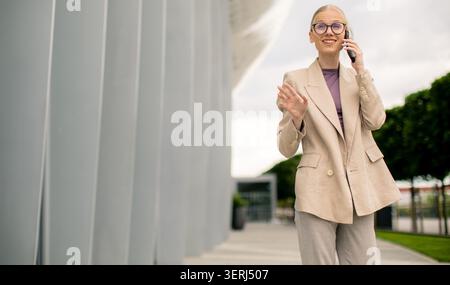 Eine Frau steht draußen und spricht mit einem Lächeln am Telefon. Sie trägt einen leichten Anzug und steht vor einem großen Gebäude mit einem modernen des Stockfoto